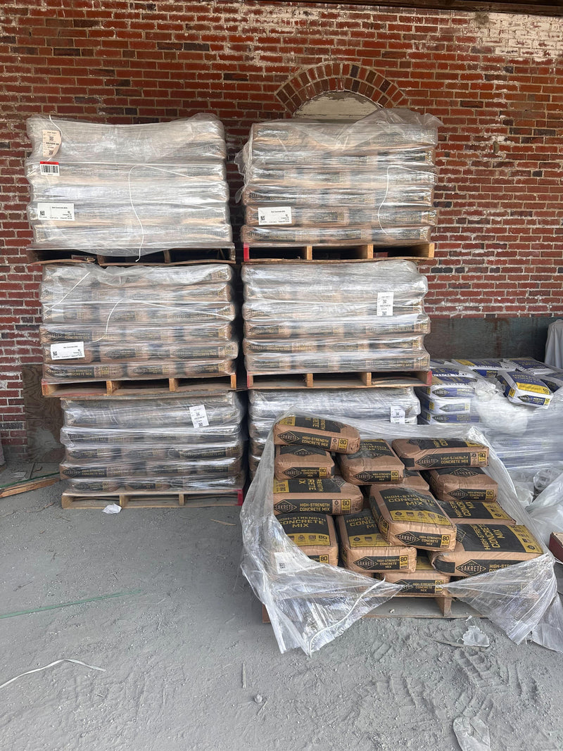 A portrait-oriented view of a selection of Sakrete Concrete products on pallets, including High-Strength Concrete Mix and Mortar/Stucco Mix Type S. Two pallets are unwrapped but still show remnants of plastic wrap, while other pallets with additional Sakrete products are visible in the background, wrapped.